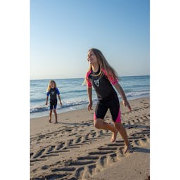 Two girls running on the beach wearing the blue and pink EVO Kid's Shorty Wetsuits Thumbnail}