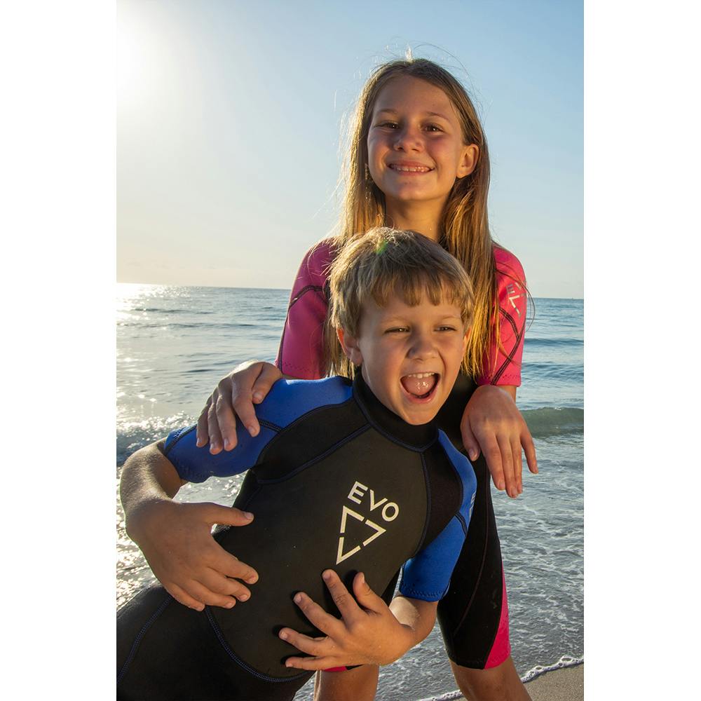 A girl and boy on the beach smiling, wearing the blue and pink EVO Kid's Shorty wetsuits